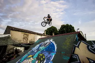 Rider casqué en BMX en saut au-dessus d'un module graffé, devant une maison, spectateurs à l'ombre
