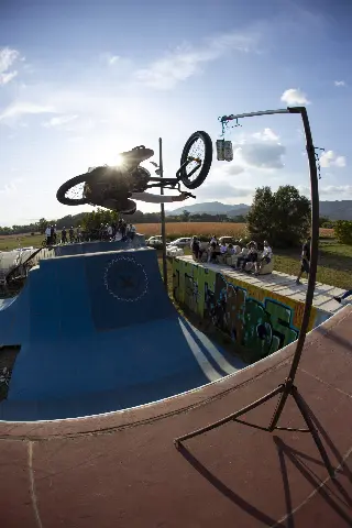 Rider en BMX en saut au-dessus d'une rampe bleue, spectateurs assis sur un module graffé, canettes suspendues à une perche