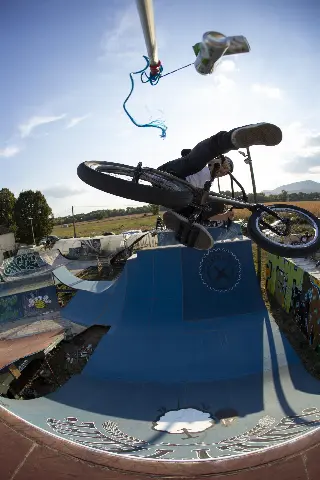 Rider en BMX en l'air sous une perche d'où pend une canette, au-dessus d'une rampe bleue