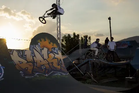 Rider en BMX en saut au-dessus d'une courbe graffée au coucher du soleil, d'autres riders attendent sur une plateforme