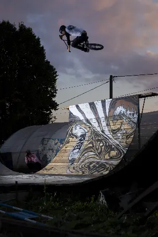 Rider en BMX en saut au-dessus d'une grande rampe en bois couverte d'une fresque, sous un ciel crépusculaire