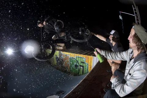 Rider en BMX de nuit au-dessus d'une rampe graffée, arrosé par deux spectateurs avec une bouteille