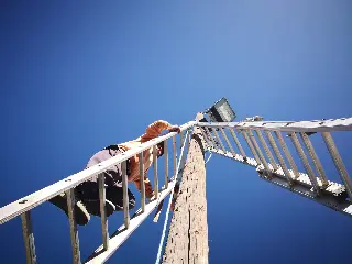 Personne sur une échelle appuyée contre un poteau en bois portant un projecteur, vue en contre-plongée
