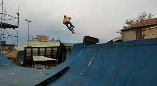 Rider en sweat orange en saut au-dessus d'une rampe bleue, échafaudage et bâtiments en arrière-plan