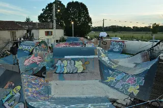 Vue d'ensemble du park graffé en fin de journée, un rider en saut au fond et des spectateurs