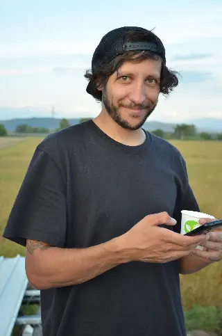 Homme souriant en casquette à l'envers, un téléphone et un gobelet à la main, champ derrière