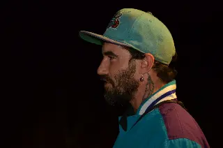 Homme barbu de profil en casquette verte et veste colorée, sur fond noir