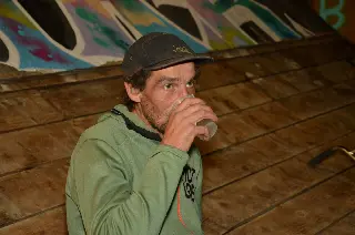 Homme en casquette et sweat vert buvant dans un gobelet, devant une rampe en bois graffée