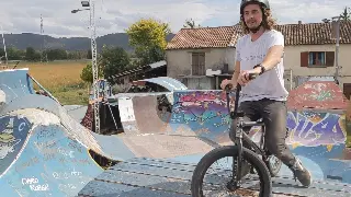 Rider casqué aux cheveux longs sur son BMX, rampes graffées et maison derrière lui