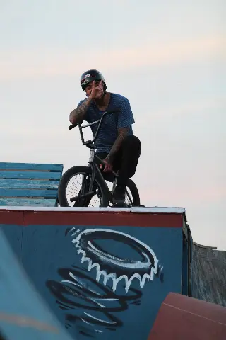 Rider casqué et tatoué assis sur son BMX en haut d'une rampe, la main devant le visage